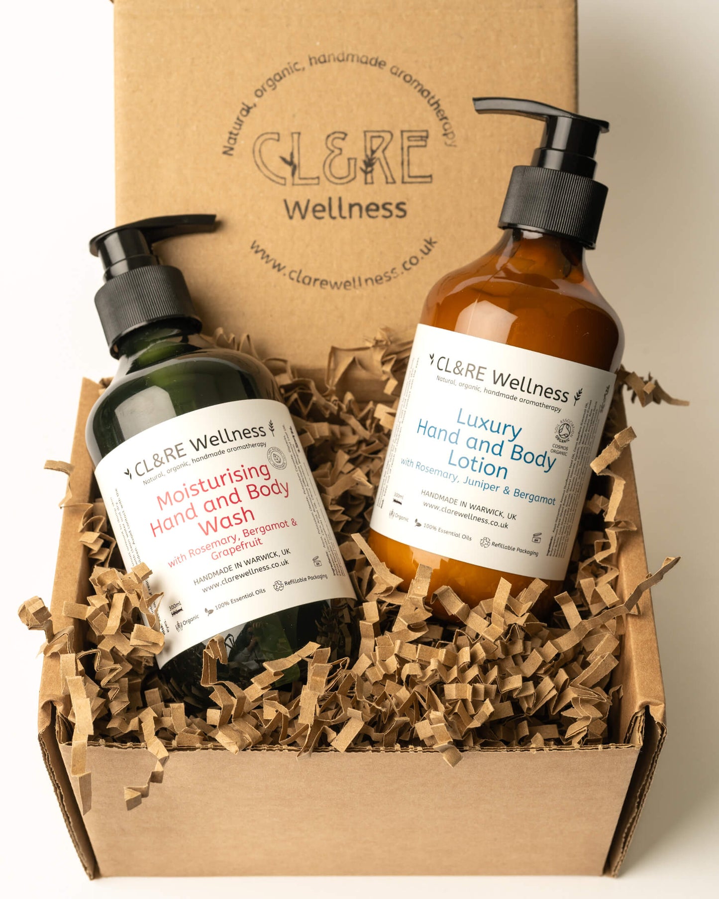 Two CL&RE Wellness hand and body products displayed in a kraft gift box with shredded paper filler: a dark green bottle of Moisturising Hand and Body Wash and an amber bottle of Luxury Hand and Body Lotion.