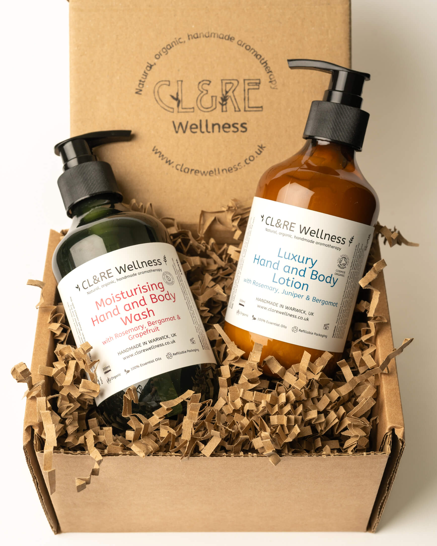 Two CL&RE Wellness hand and body products displayed in a kraft gift box with shredded paper filler: a dark green bottle of Moisturising Hand and Body Wash and an amber bottle of Luxury Hand and Body Lotion.