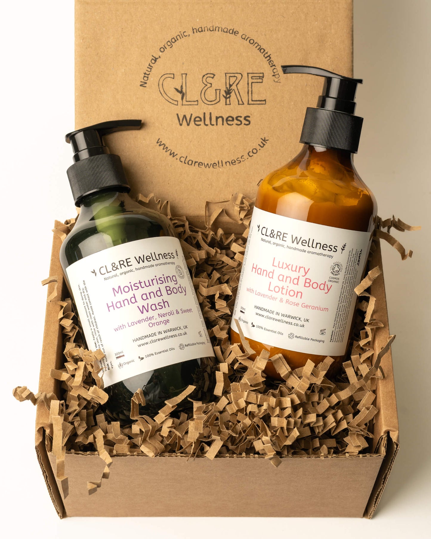 A CL&RE Wellness gift set containing a green bottle of Moisturising Hand and Body Wash and an amber bottle of Luxury Hand and Body Lotion, both with black pumps, nestled in brown paper shred inside a cardboard box with the branded inner lid behind them.