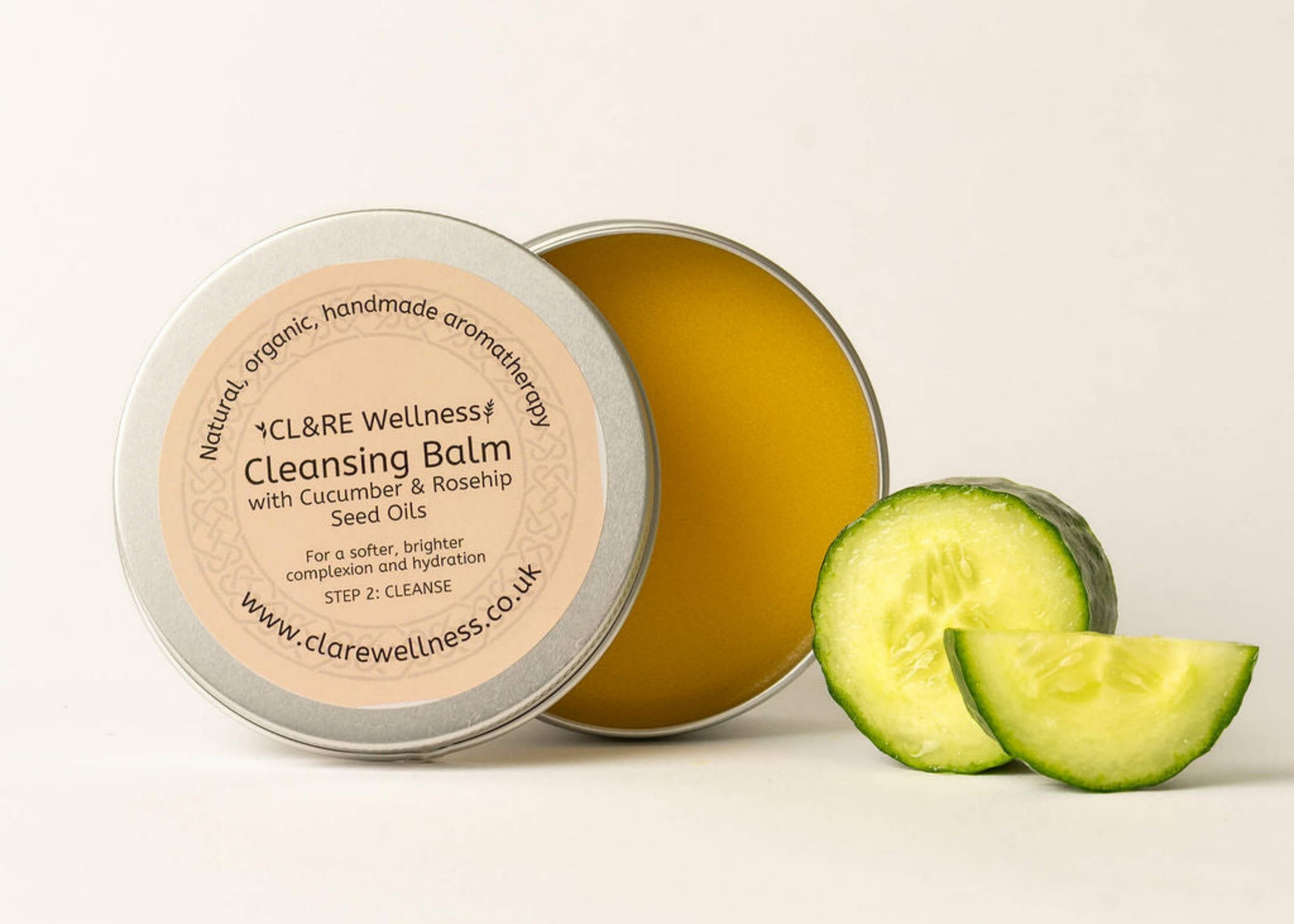 An open container of cleansing balm with a label that reads 'Cleansing Balm with Cucumber & Rosehip Seed Oils'. The container is accompanied by slices of cucumber next to it.