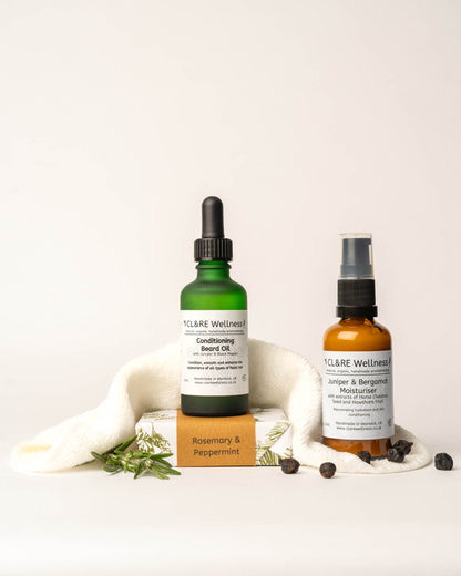A Conditioning Beard Oil bottle and Facial Moisturiser bottle from CL&RE Wellness on a light background with a wash cloth, rosemary sprigs and juniper berries.