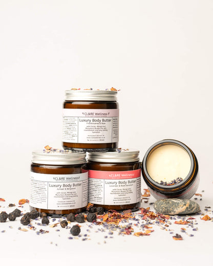Four jars of CL&RE Wellness luxury body butter with visible branding on a white background and scattered botanicals around them.