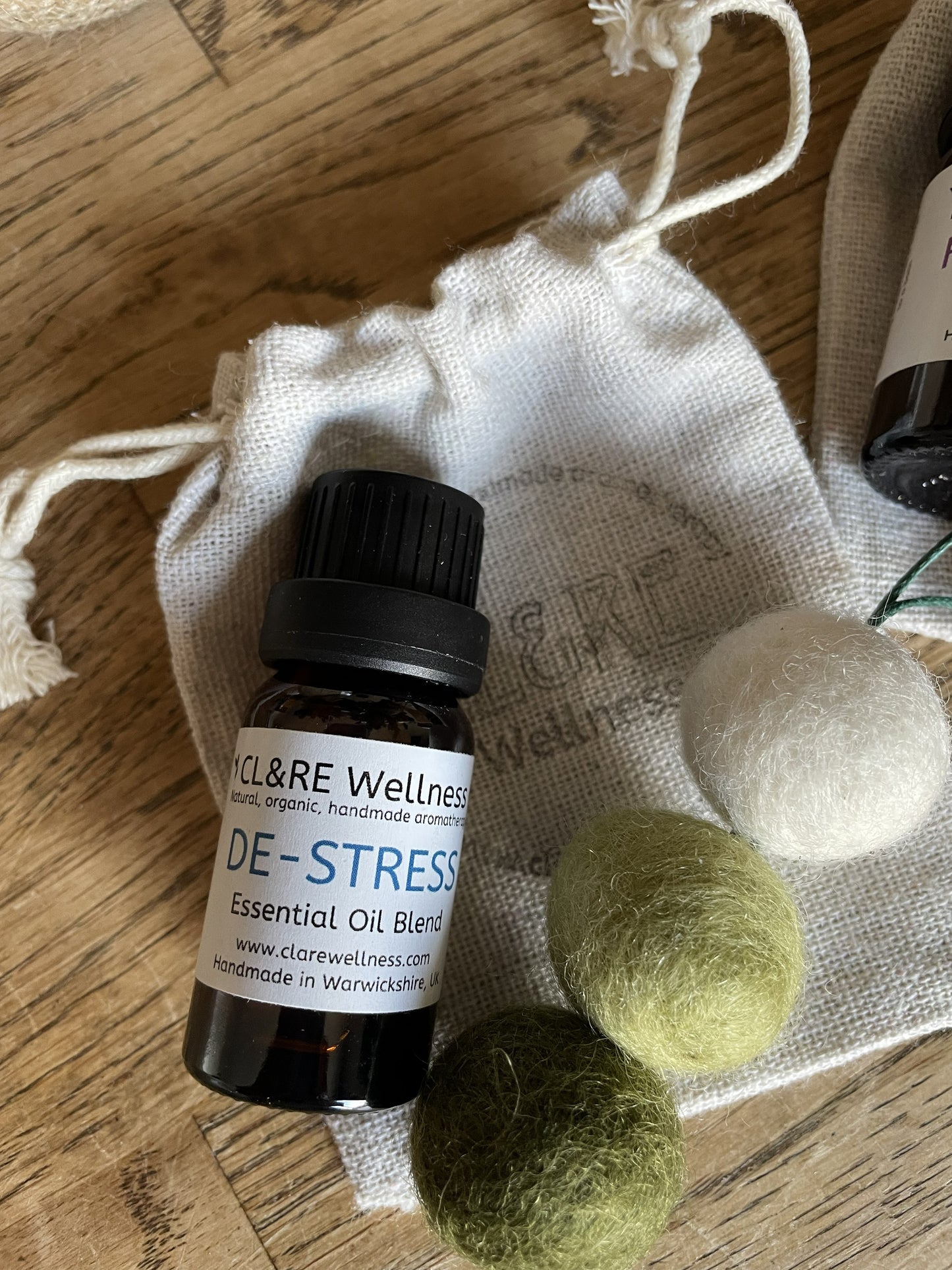 Bottle of CL&RE Wellness DeStress Essential Oil blend with a green coloured natural wool felt diffuser and a linen bag, on a wooden table.
