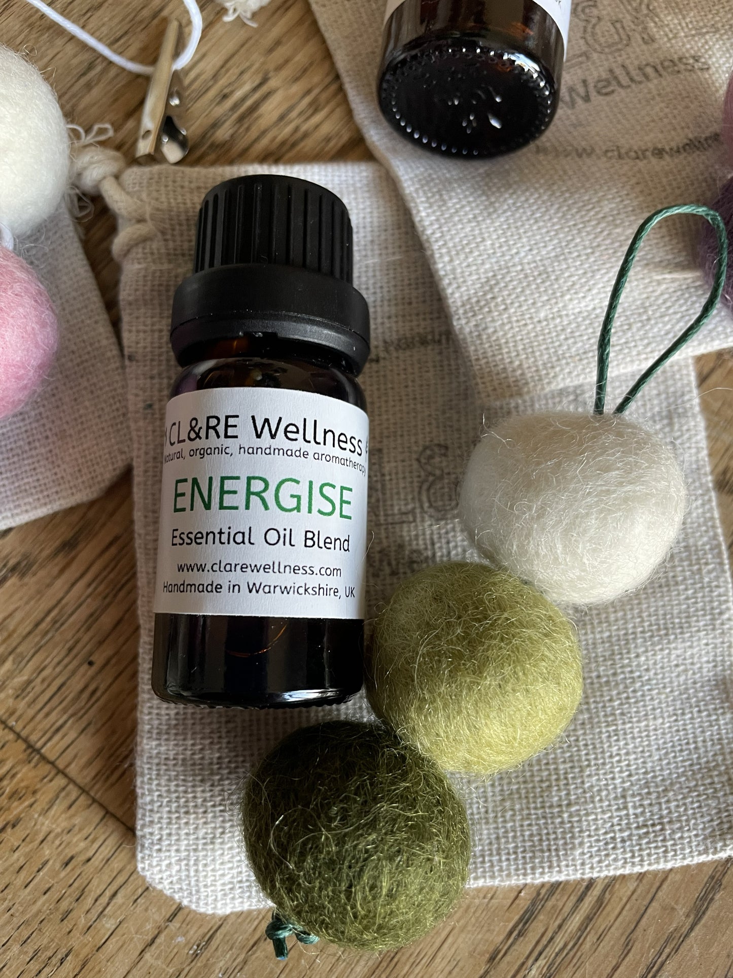 Bottle of CL&RE Wellness Energise Essential Oil blend with a green coloured natural wool felt diffuser and a linen bag, on a wooden table.