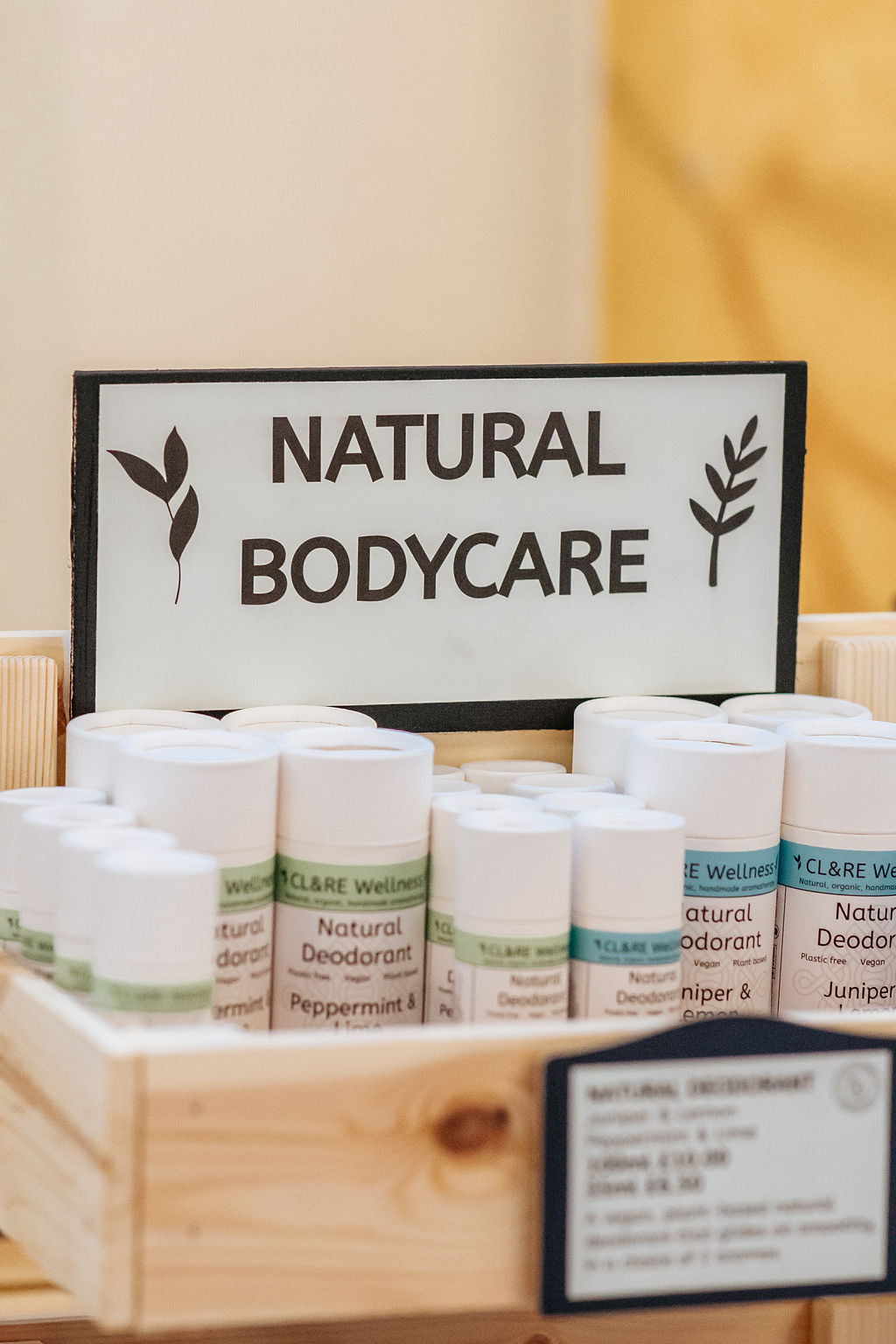 A row of natural deodorants in white cardboard tubes with CL&RE Wellness branded label saying Natural Deodorant Juniper & Lemon, Plastic-free, Vegan and Plant Based, in a wooden rack with a sign Natural Bodycare.