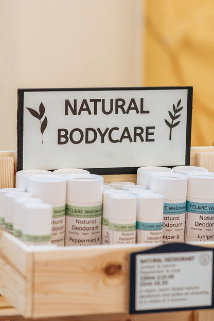 A row of natural deodorants in white cardboard tubes with CL&RE Wellness branded label saying Natural Deodorant Juniper & Lemon, Plastic-free, Vegan and Plant Based, in a wooden rack with a sign Natural Bodycare.
