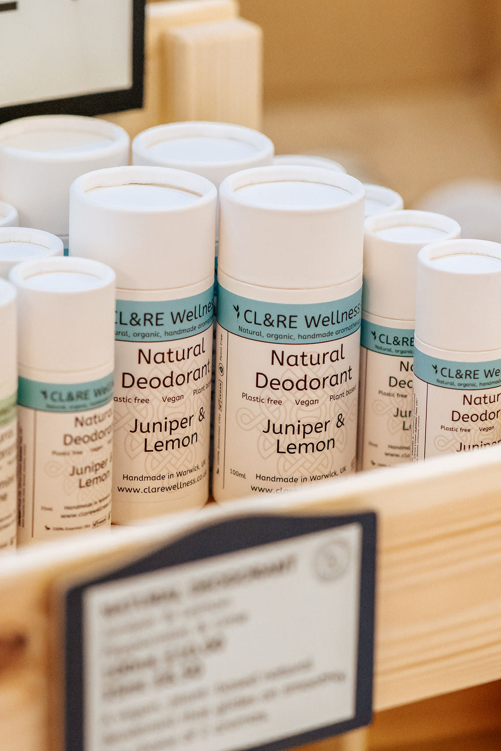 A row of natural deodorants in white cardboard tubes with CL&RE Wellness branded label saying Natural Deodorant Juniper & Lemon, Plastic-free, Vegan and Plant Based, in a wooden rack.
