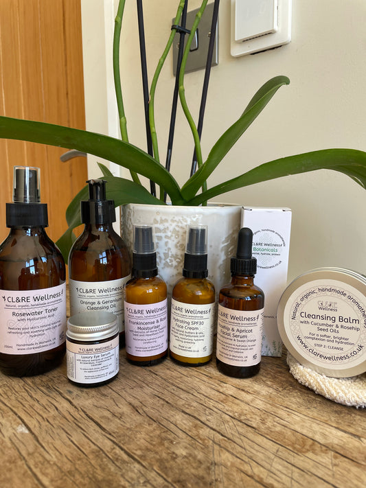 A collection of skincare products from CL&RE Wellness arranged on a wooden table in front of a potted plant. The products include amber spray and dropper bottles, a small jar, a white box, and a round tin of cleansing balm.