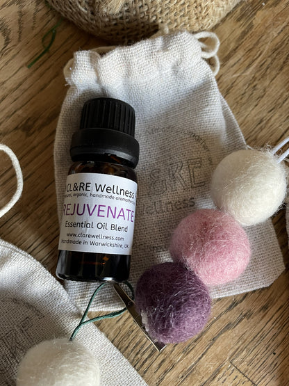 Bottle of CL&RE Wellness Rejuvenate Essential Oil blend with a purple colored natural wool felt diffuser and a linen bag, on a wooden table.